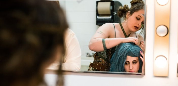 One actress helping another actress with her wig in a mirror backstage