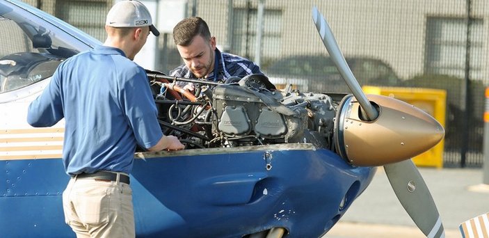 The nose of a plane with the engine exposed and two people working on it