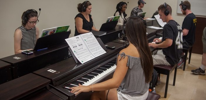 Eight pianos back to back down the middle of a classroom with a student playing and wearing headphones at each one.