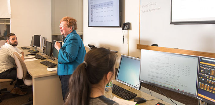Professor Marian Extejt teaching a class standing at the front of the classroom flanked by two tables each with a student sitting at a computer