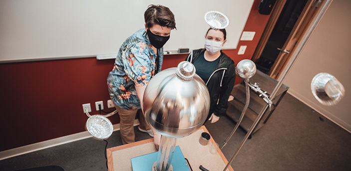 2 BSU students operating an electro magnetic tower with small foil pie plates on the top and flying in the air around it