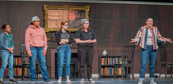 5 actors on a stage with a backdrop of a fireplace and bookcases. One actor is separate from the others, speaking and gesturing while the others look on.