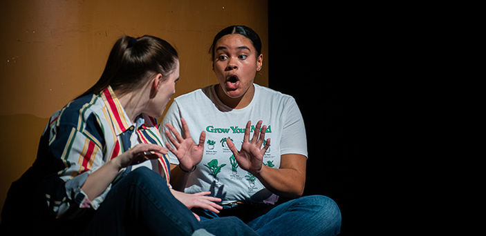 2 actors sitting on stage looking at each other and talking with one holding her hands up and her mouth open.