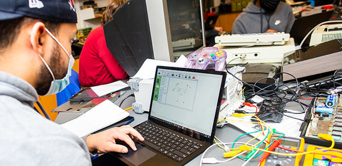 3 students in computer class working on laptops and sitting at a table covered with an old tv screen, video game controllers, computer boards and other electronic equipment