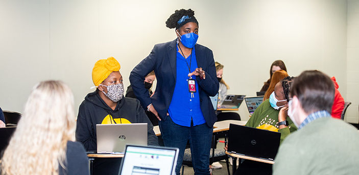 Professor Castagna Lacet stands talking to a circle of students sitting in desks working as a group with laptops open with another circle of students working in the background