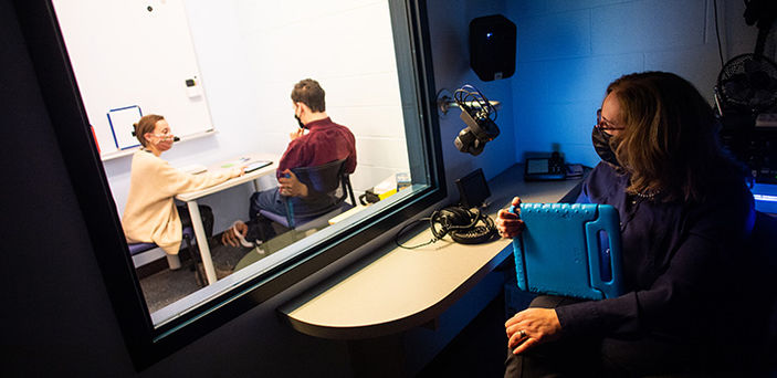 A BSU student works with a young man in the BSU Speech Language Literacy Center while a professor watches through one way glass