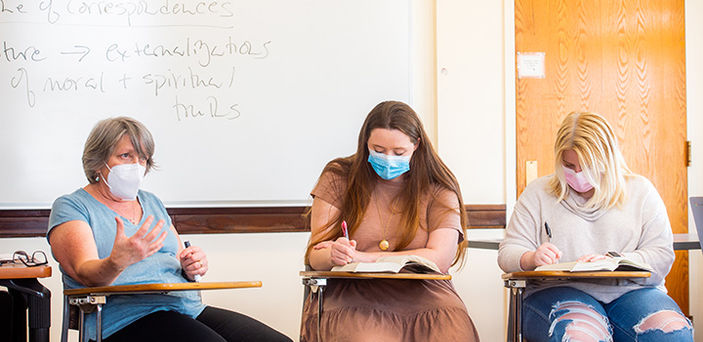 Dr. Ann Brunjes sits at a desk teaching in front of a white board that shows partial notes that read "correspondences" and " externalizations of moral and spiritual truths." Two students sit at desks beside her taking notes in their books.