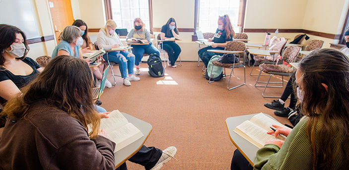 Dr. Ann Brunjes and a circle of students sitting in desks with books open and pens out discuss the book they are reading