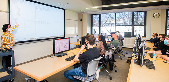 Dr. Enping Li teaching Computer Forensics class pointing at the projector screen while students sitting in rows of tables with computers look on