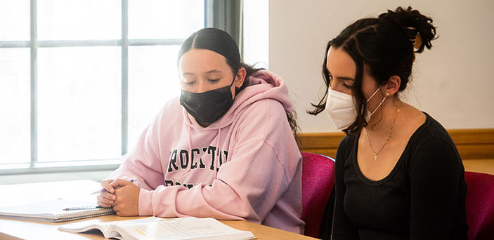 2 students sit at a desk in class looking at a book with notebook and pens around it