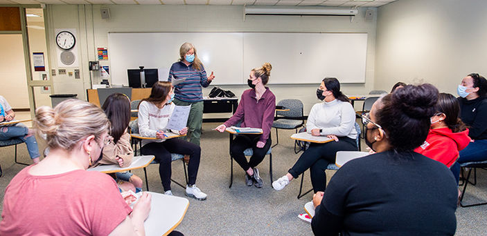 students sit in a circle in individual desks listening to Professor Leslie Sattler who stands talking to one of the students