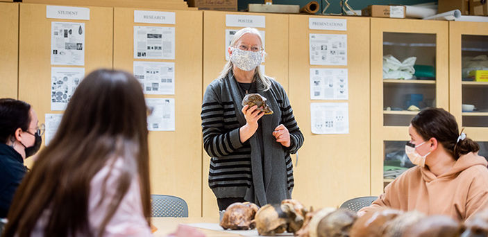 BSU Professor Ellen Ingmanson holds a skull while she teaches at the head of a table with a row of skulls on it that students sit around listening and taking notes