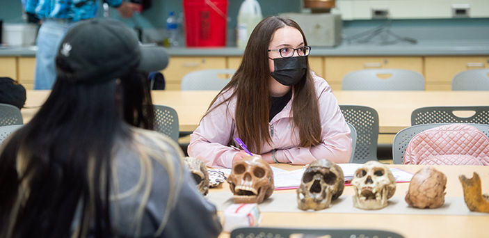 2 students sit at a table in front of a row of skulls listening to the professor and taking notes
