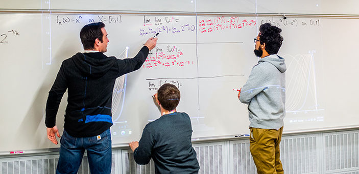Professor Matt Salomone points to part of an equation written on a white board by a student who stands with a marker in his hand listening to the professor while another student works on an equation on the board
