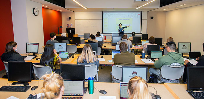 view from the back of the room of Dr. Haleh Khojasteh's Computer Science I class where she points at content on a projector screen in front of rows of students at desks with laptops open 