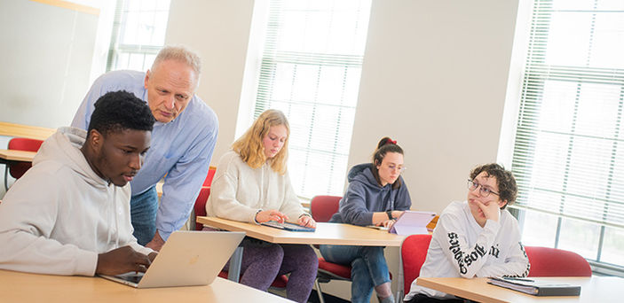 Professor Joseph D'Adamo helps a student in class, leaning down to look at the student's open laptop with him