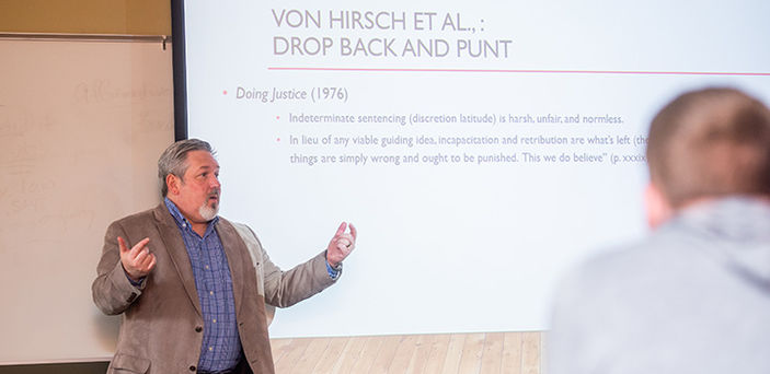 Professor Michael Devalve teaching in front of a projector screen with information from Doing Justice (1976)