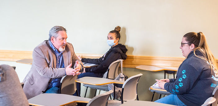 Professor Michael Devalve sitting in a classroom desk talking with a student in class