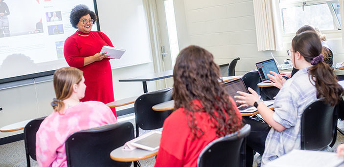 BSU Professor Sheena Manuel standing in front of a projector screen smiling and holding a notebook while listening to a student speaking in class