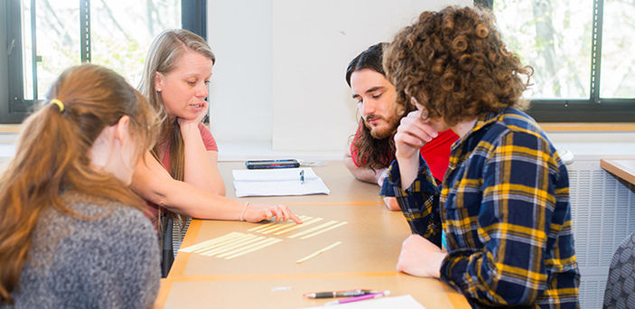 Professor Shannon Lockard sits at a classroom table with 3 students explaining something with strips of paper laid out on the table