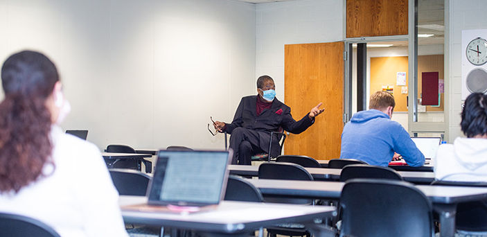 BSU Professor Walter Harper sitting and teaching at the front of the classroom while students sit at rows of tables with laptops open