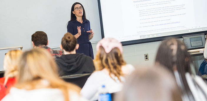 BSU Professor Ashley Rodrigues standing next to a projector screen teaching as students look on