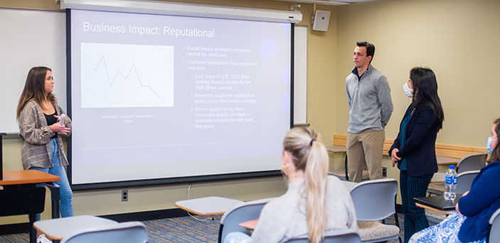 Professor Hui Zhang talks to 2 students giving a presentation in a Public Relations class with a projector screen titled "Business Impact: Reputational" at front center