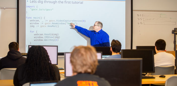A faculty member teaches pointing to a projector screen as students with open laptops look on