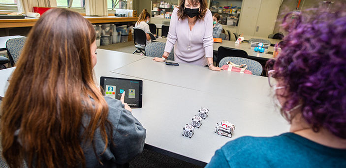 Professor Jeanne Ingle leans over a classroom table while 2 students sitting at the table work with an ipad and electronic blocks and vehicle