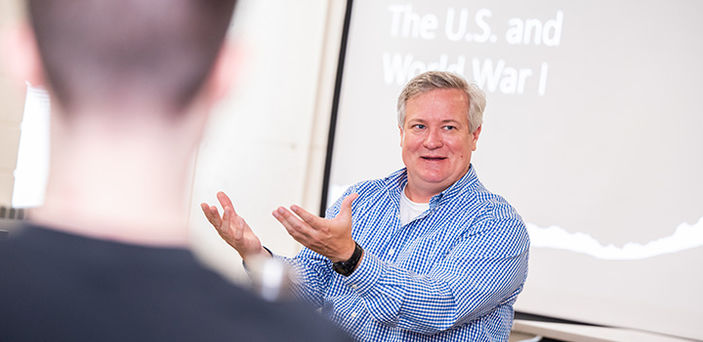 Professor Thomas Nester teaching American Military History sitting in front of a projector screen that says "The U.S. and World War I"