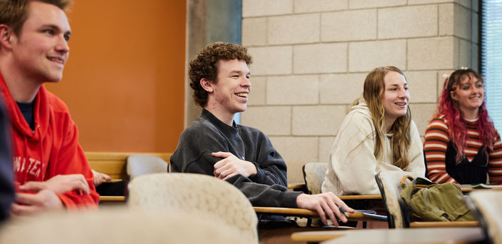 4 students sit in a row of individual desks smiling at the teacher