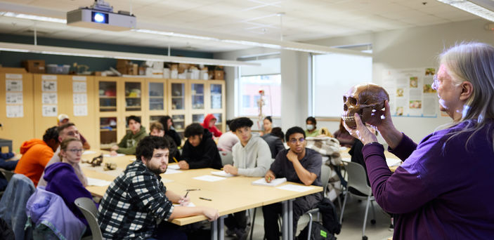 Dr. Ellen Ingmanson holds a skull while students sitting at 2 large tables in class look on