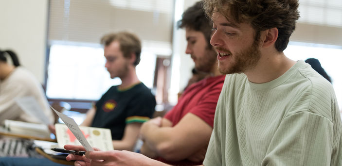 Students sit in a row of individual desks while the student closest to the camera reads from a paper with pen in hand