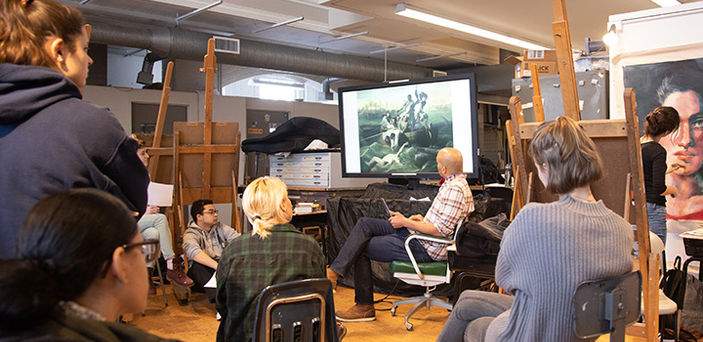 a professor and small group of students view paintings on a large screen while a student outside the group paints a large portrait