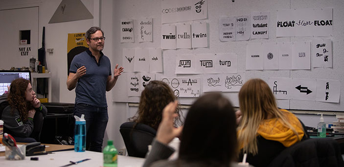 A BSU professor speaks at the front of a graphic design class beside a wall covered with graphic depictions of words