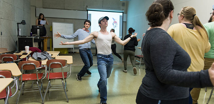 an acting class with the students walking in a circle with their arms out wide; one looks like he's walking on a catwalk or runway while others walk with looser posture