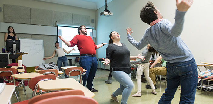an acting class with the students walking in a circle with their arms out wide, leaning back and yelling while the teacher directs from the front corner of the room