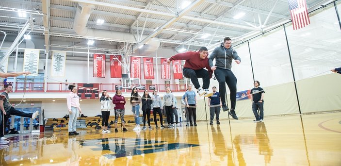 2 male students jumping over a rope in Physical Education class in a gymnasium as other students cheer them on