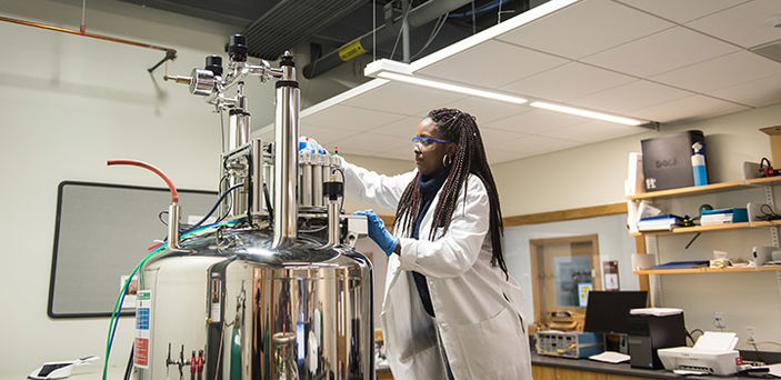 a female BSU student wearing lab coat, rubber gloves and safety glasses stands on a step stool to reach the top of a large silver piece of equipment