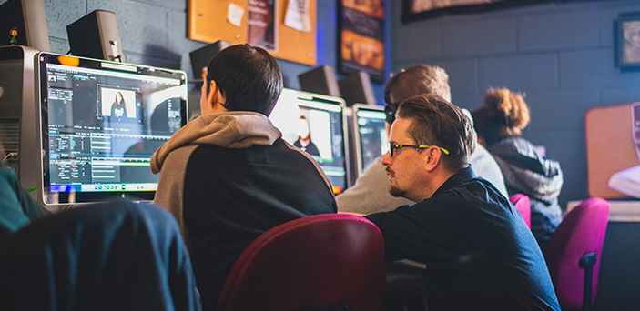 a professor helps a student in the BSU Communication Studies Editing Lab where a row of students sit at computers editing video