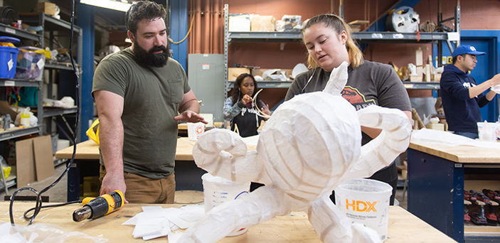 BSU student and woodworking professor discuss the student's project, a wooden framed octopus covered in white fabric, while 2 other students work in the background