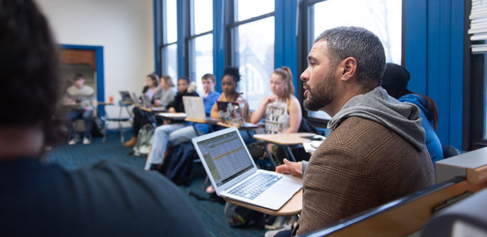 Professor Justin Caouette teaching with his laptop open sitting in a desk in a circle of students at desks with their laptops open. 