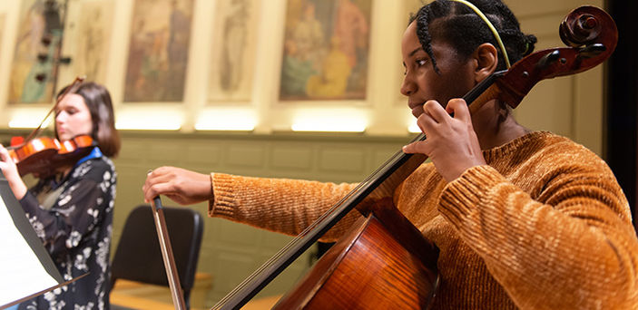 a BSU student in foreground playing the cello and another student in the background playing violin