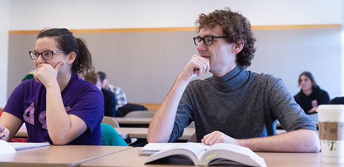 2 students sitting at a classroom desk smiling while listening to the professor