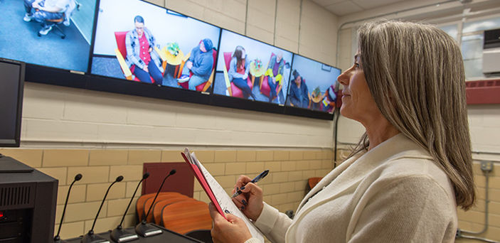 Dr. Christy Lyons Graham watching students' practice counseling sessions on a row of TV monitors and taking notes