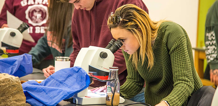 A BSU student in Forensic Geology class looking into a microscope with a rock on the table behind the microscope and 3 other students in background working at the table