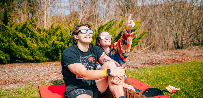 Two students sitting on a blanket gaze and point at the sun while wearing glasses.