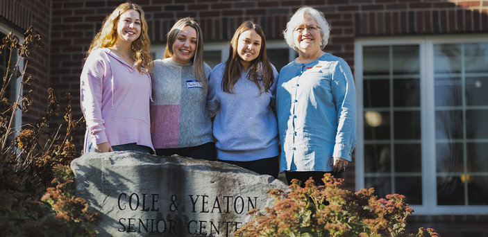 Three students and their professor stand behind a senior center sign.