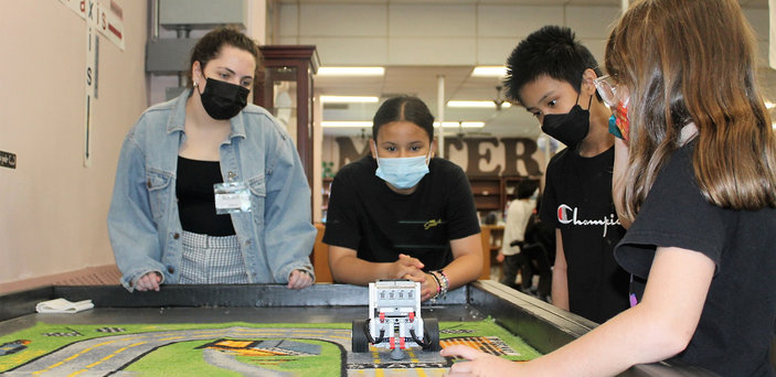 A BSU student helps fifth graders test a robot on a mat.