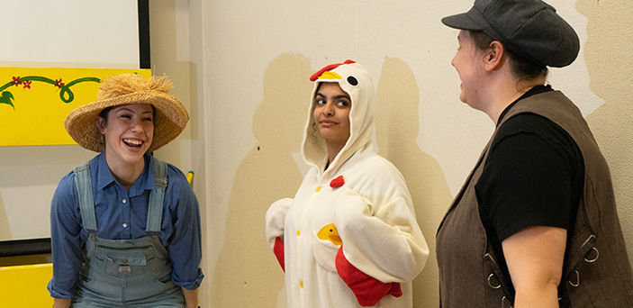 2 students playing farmers stand laughing on either side of a student playing a chicken